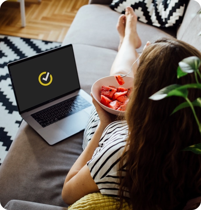 Kvinde sidder på en sofa og spiser vandmelon, mens hun åbner Norton 360 AntiVirus Plus på sin laptop